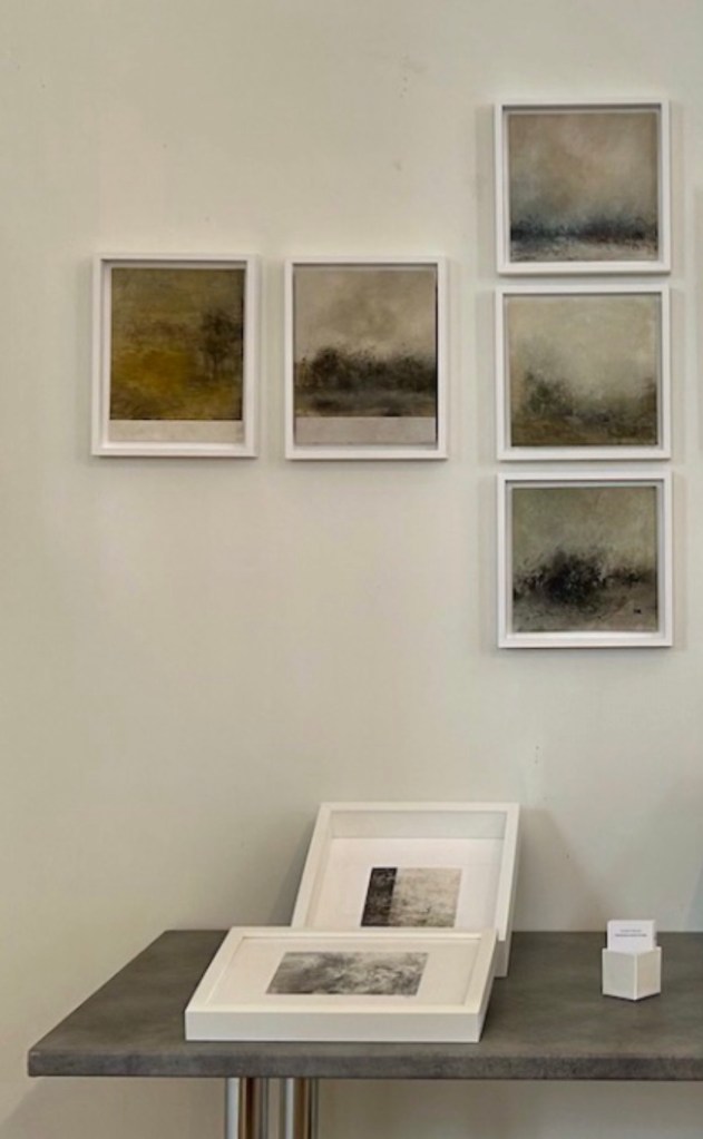 A collection of framed abstract artworks displayed on a wall, with a small table in front holding additional framed pieces and a small box.